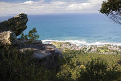 This image captures a scenic view from a high vantage point, likely a cliff or hill, overlooking a coastal town. Large rock formations and vegetation, such as bushes and small trees, are in the foreground. Below, there is a densely forested area leading to a small town with houses and buildings. The vast ocean stretches beyond the town, with waves breaking on the shore. The sky is partly cloudy with blue patches visible.