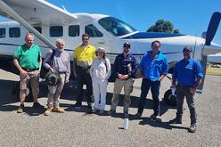 A group of seven individuals standing in front of a white propeller aircraft with blue and black detailing. The individuals appear to be pilots or aviation staff, indicated by their uniforms and headgear. The setting is outdoors on a clear, sunny day.