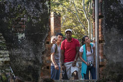 Four people with obscured faces standing amidst the ruins of a brick structure with overgrown vegetation, suggesting a scene of exploration or discovery in an abandoned location.