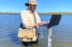 An individual stands in a body of water while using a laptop, with a measuring stick indicating the water depth. The person is wearing a wide-brimmed hat and carrying a shoulder bag, possibly for work-related items. This image highlights an unconventional work setting, possibly for remote work or environmental research.