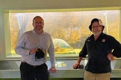 Two individuals standing in front of a large aquarium tank with various fish and aquatic plants visible inside.