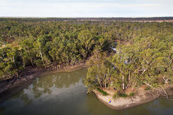 An aerial photo of Sandy Bend at Woolpress Bend campground in Yanga National Park. The image shows a river bend surrounded by lush vegetation, under a bright blue sky with scattered clouds.