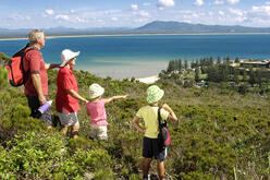 A group of four people, consisting of two adults and two children, are standing on a grassy hill overlooking a scenic coastal landscape. The adults are wearing hats and carrying backpacks, while the children are wearing sun hats. One of the children is pointing towards the ocean. The background features a beautiful view of the ocean with clear blue water, a sandy beach, and a distant mountain range under a partly cloudy sky. The scene is serene and picturesque, capturing a moment of outdoor exploration and 