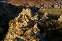 Panoramic view of the Gardens of Stone reserves, featuring rocky mountains of varying heights, grassy hills and shady valleys. There are two people hiking on one of the rocky hills carrying red backpacks and wearing hiking clothes. The sun illuminates some of the mountains, while casting shade across the valley.