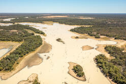 Aerial photograph of a still, sand-coloured river on a long, endlessly flat plain of sand-coloured earth and dark vegetation